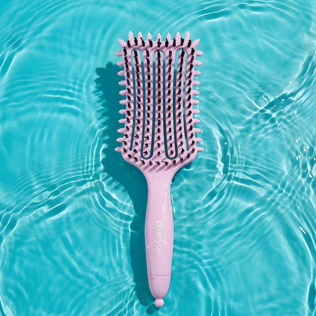 PixieYou Cloud Brush in pink, featuring a wide paddle design, resting on a turquoise water surface.