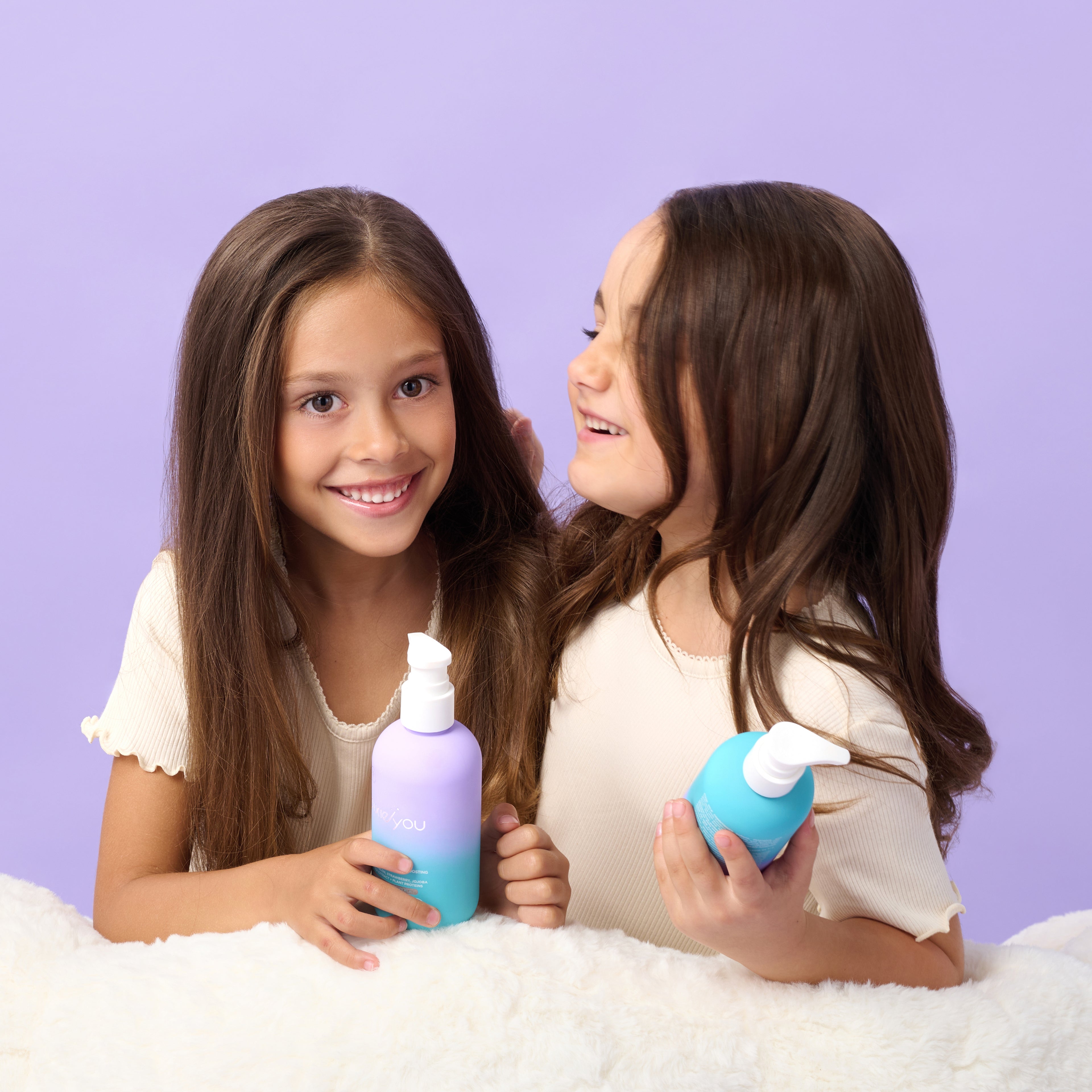 Two smiling girls hold PixieYou shampoo bottles against a soft purple background, showcasing the product's vibrant packaging.